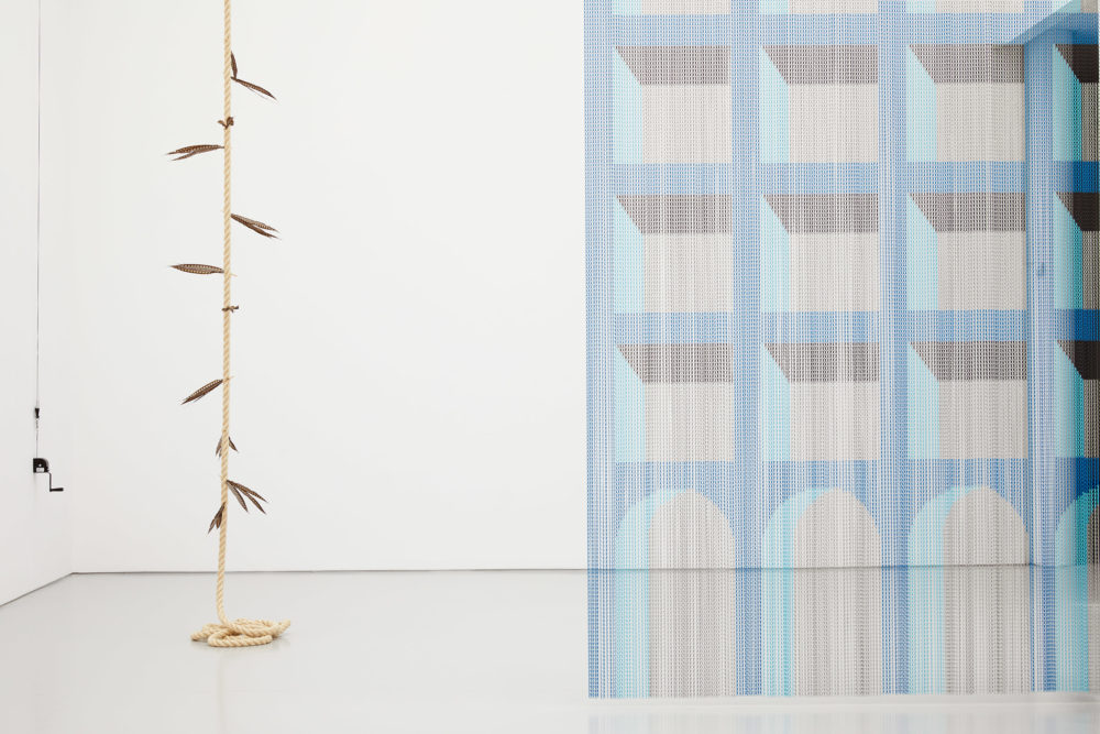 A curtain made from blue beads arranged to look like windows and doors in a large building. To the left is a rope hanging from the ceiling with feathers stuck in the length.