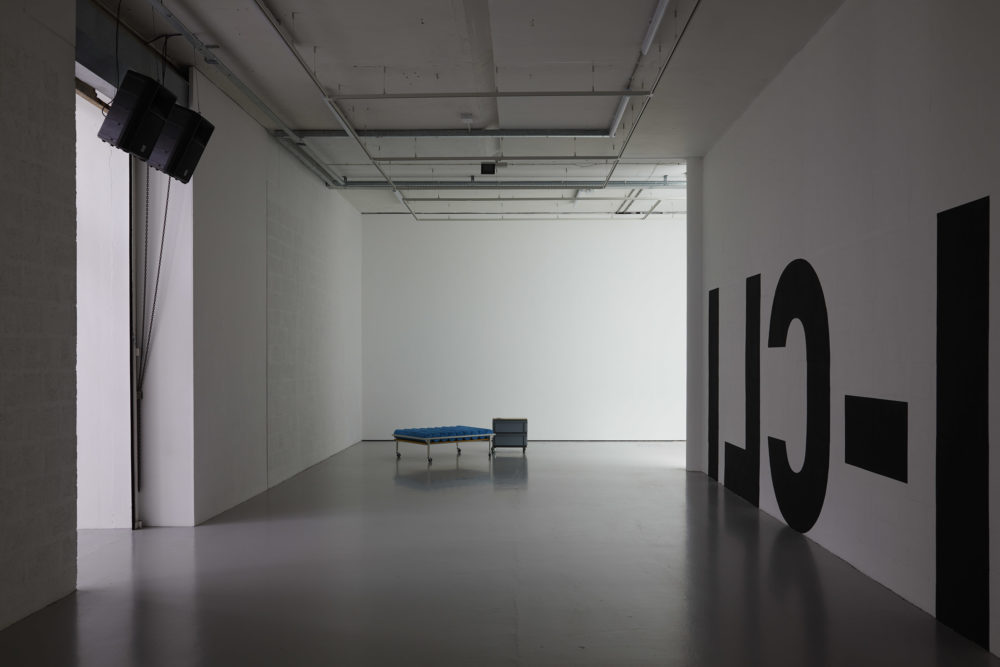 A corridor - on the right wall are large black letters written backwards. On the left wall are two large speakers. Directly in front is a medical-looking bench with a filing cabinet next to it.