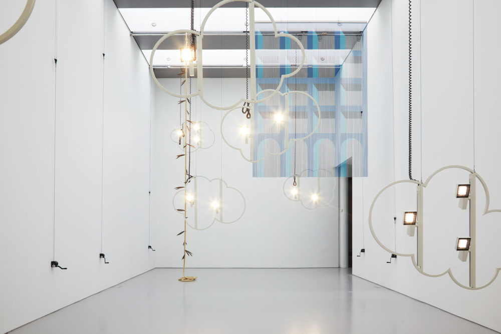 Structures outlining the shape of clouds are hung from the ceiling. Also hung from the ceiling is a bead curtain decorated to look like the side of a large building with multiple windows close together.