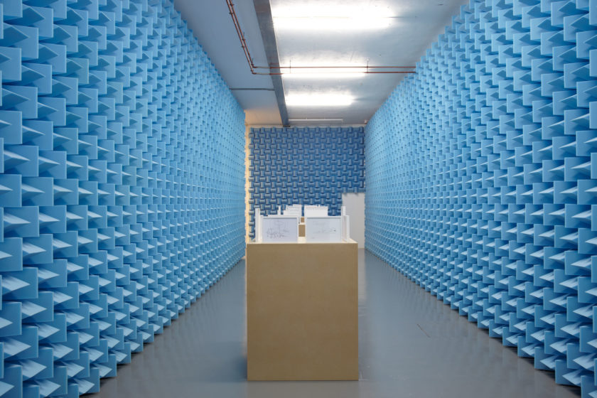 Installation view of Haroon Mirza. Small framed diagrams sit on wooden plinths in between walls covered in blue sound proofing foam.