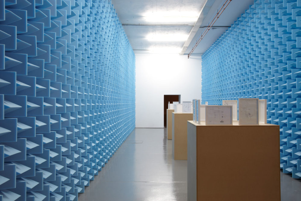 Installation view of Haroon Mirza. Small framed diagrams sit on wooden plinths in between walls covered in blue sound proofing foam.