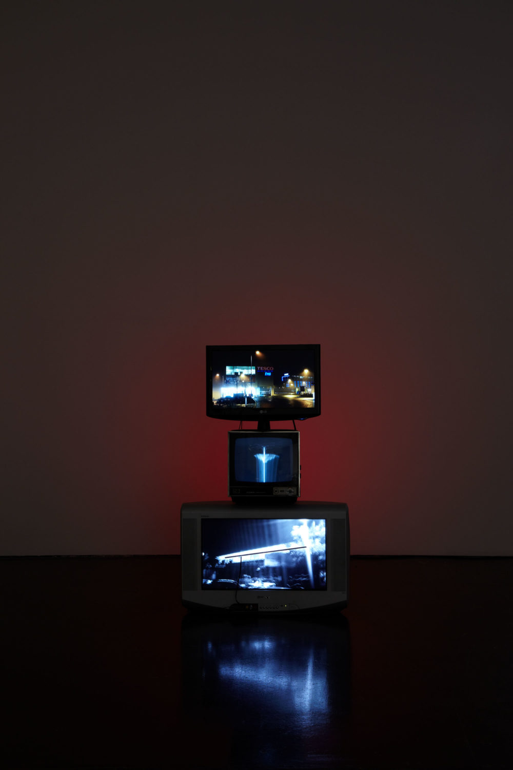 Installation view of Haroon Mirza. Three televison screens showing night scenes are stacked atop one another in a dark room.