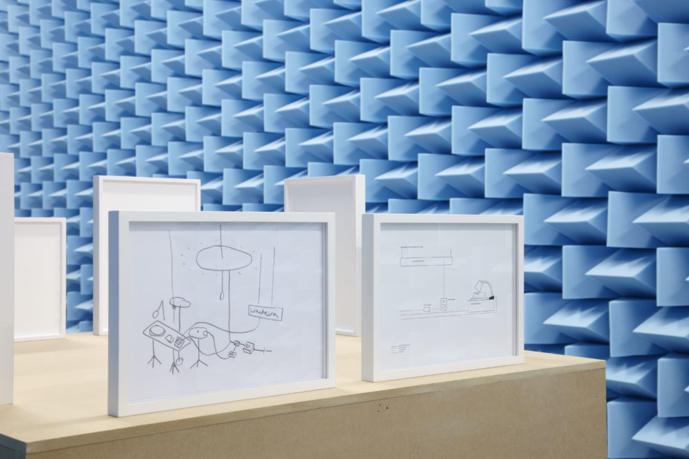 Installation view of Haroon Mirza. Small framed diagrams sit next to a wall covered in blue sound proofing foam.