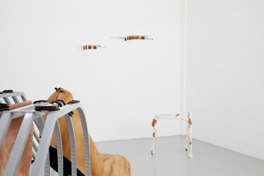 Installation: A metal frame used for displaying cars at show rooms shows in the foreground, horseshoes are placed on top of it. In the corner is a white walking frame with what looks like clay stuck to it. Coming out from the wall above are metal rails that have leather keyrings dangling from them.