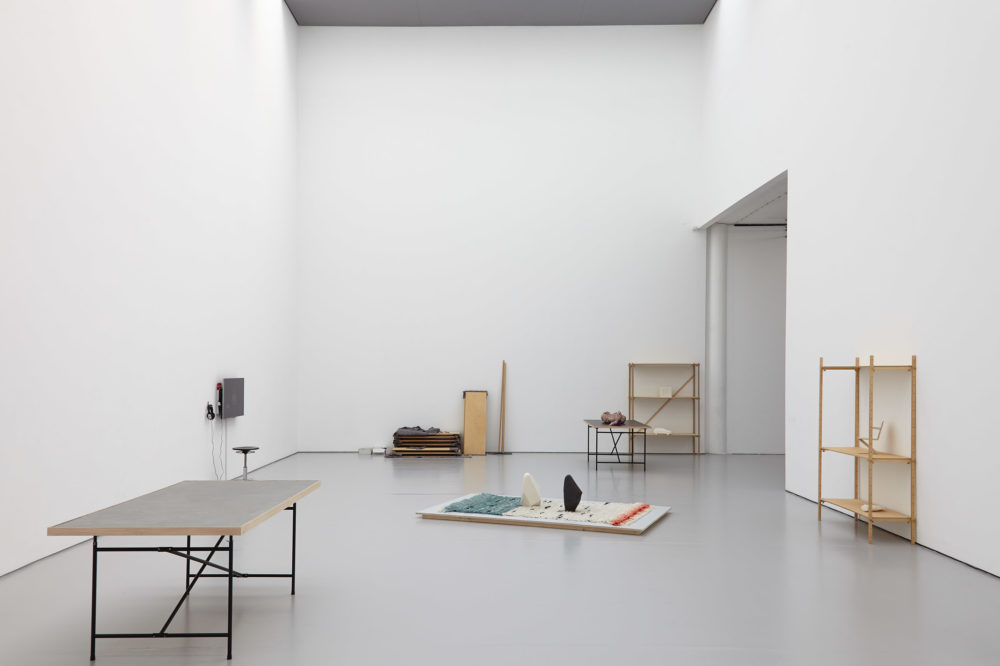 Various shelving units are place around the gallery giving an impression of a work in progress. In the middle of the gallery, on the floor is a rug with two vertical standing rocks on it.