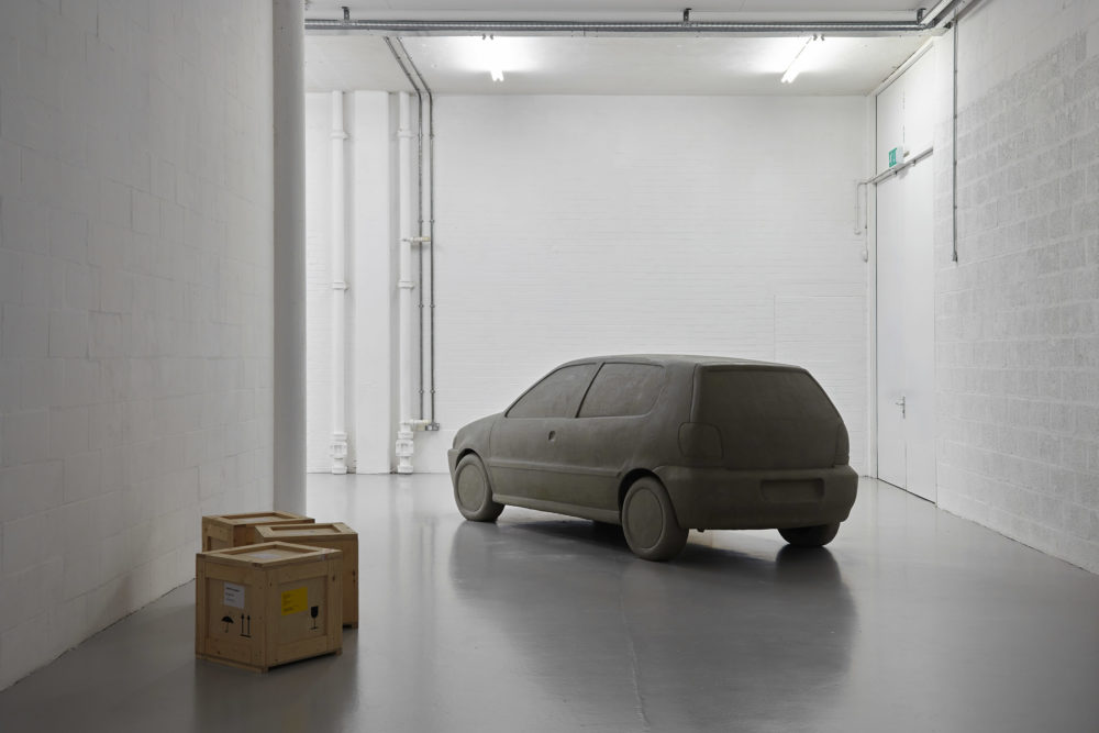 The bright white gallery has three wooden crates on the floor and a life-size plasticine car next to it.