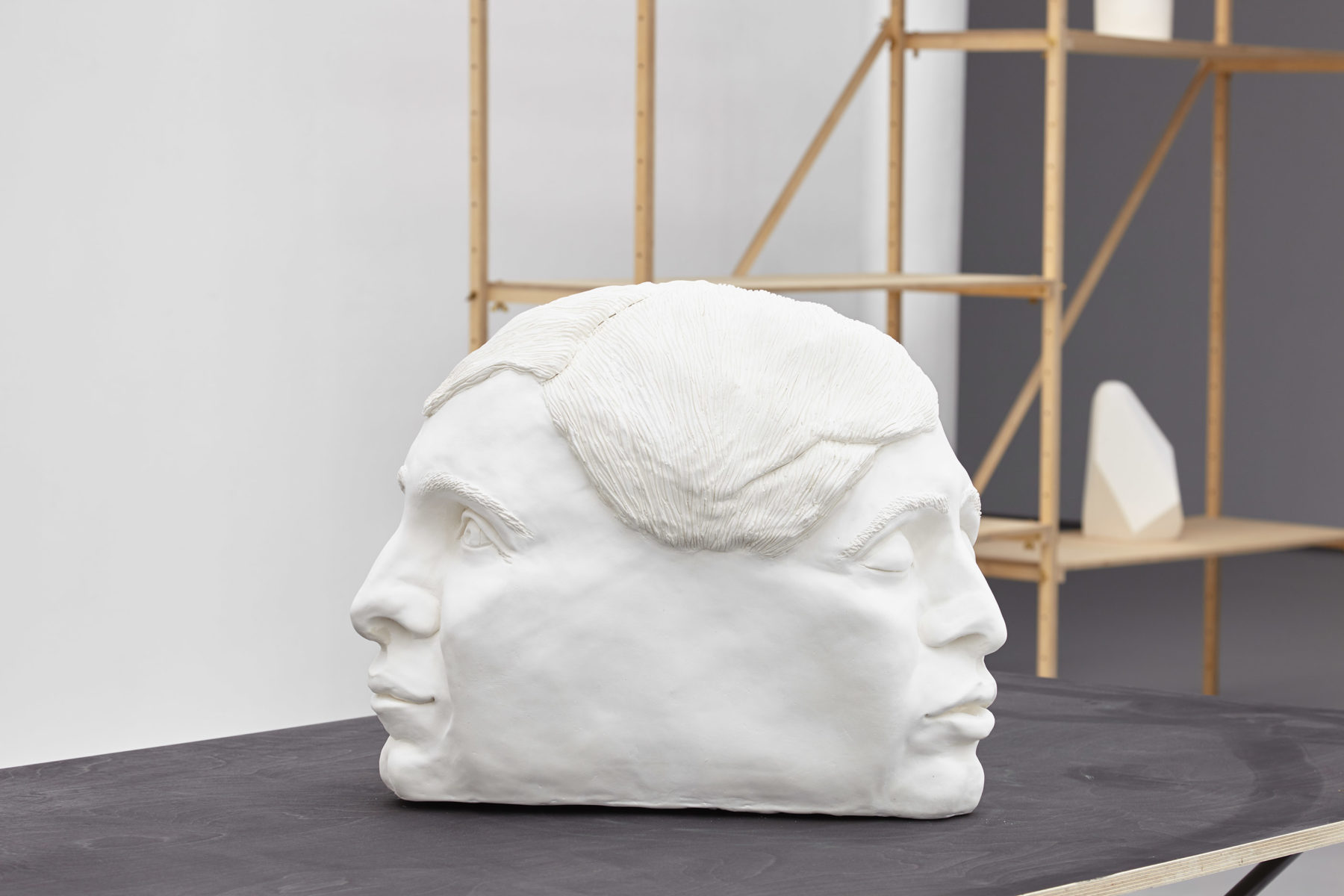 A large white sculpture of two heads joined together at the back is placed on a table in the gallery.