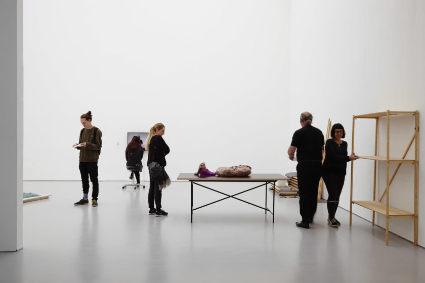 Visitors stand amongst the pieces. You can see an empty wooden shelving unit, a table with a pink clay mass on it, and on the far wall, a screen.