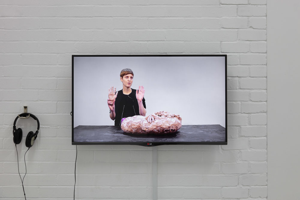 A screen in the gallery shows a person stood at a table, facing the camera. They are holding a fake hand in their right hand, and holding that up to camera alongside their own left hand. On the table before them is a large mass - perhaps of clay, that looks oddly fleshy.