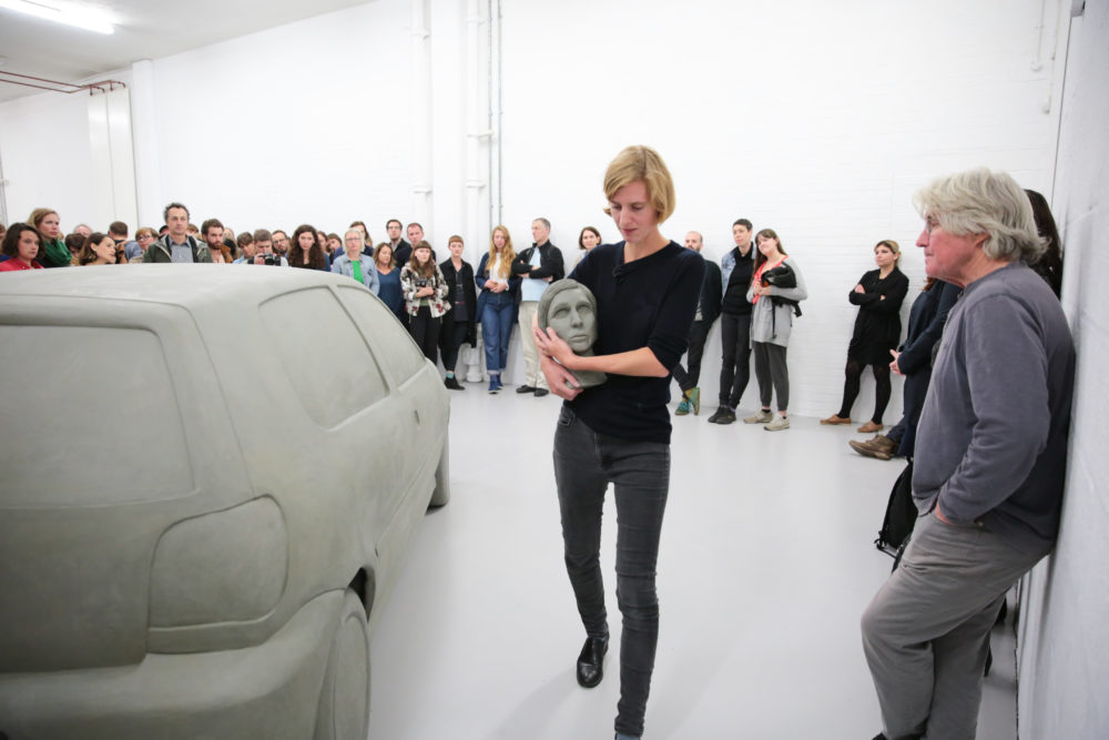 The artist holds a clay model of her own head her arms as she walks around a clay scale model of a car. This is a performance in a bright white gallery, a lot of people are standing in a large circle around her and the clay car, watching her.