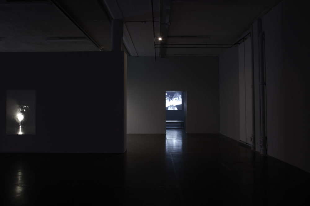 Installation shot: The gallery is dark, to the left a projector screens an image of a firework, directly in-front you can see through to another room with more video works.