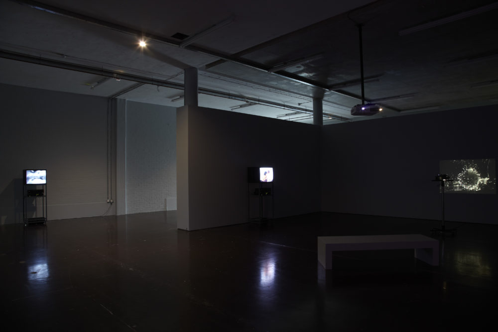 Installation shot: The gallery is dark, to the right a projector screens an image of a firework, a white bench is visible in the foreground. Two other monitors are visible.