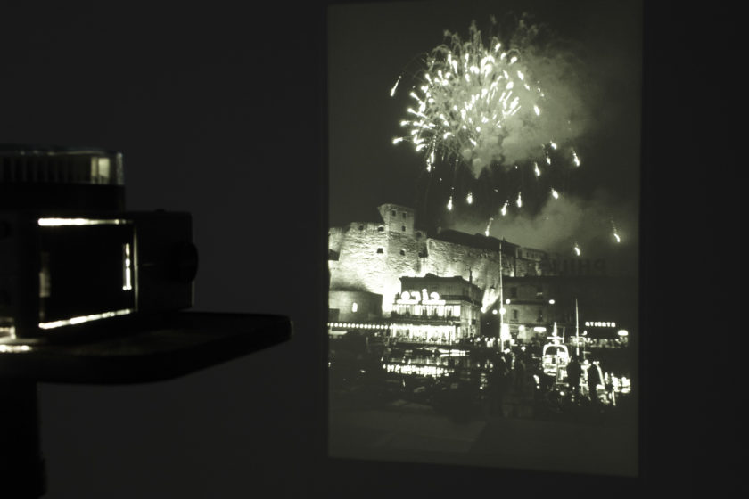 Installation shot: The gallery is dark, a projector screens an image of a firework exploding in the sky over a castle in black and white.