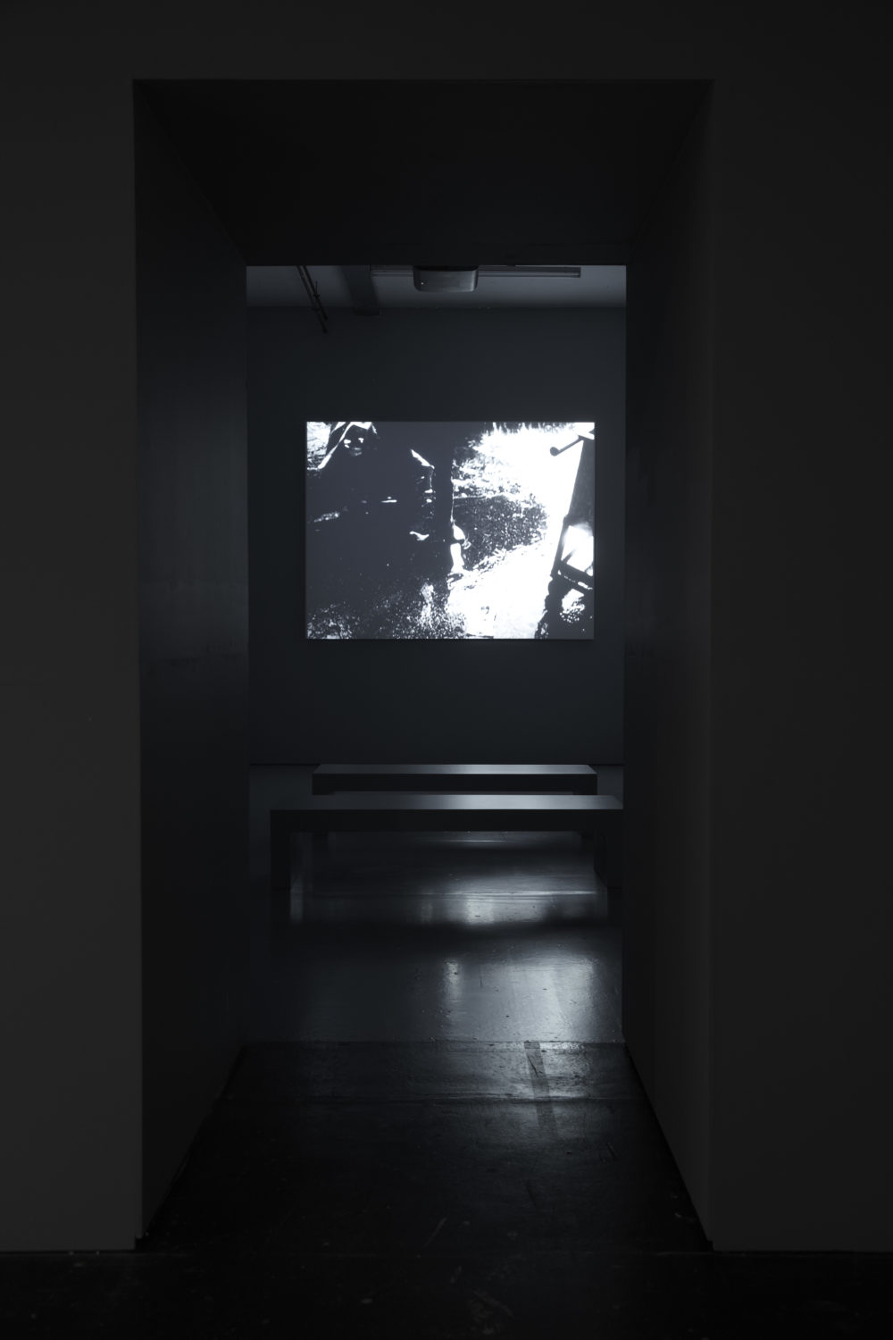 Installation shot: The gallery is dark, a screen directly in front shows art work that looks like an old black and white photograph.