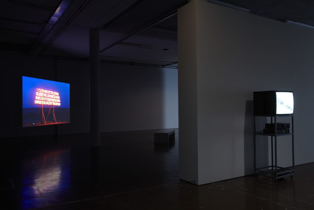 Installation shot: The gallery is dark, two screens show art work.