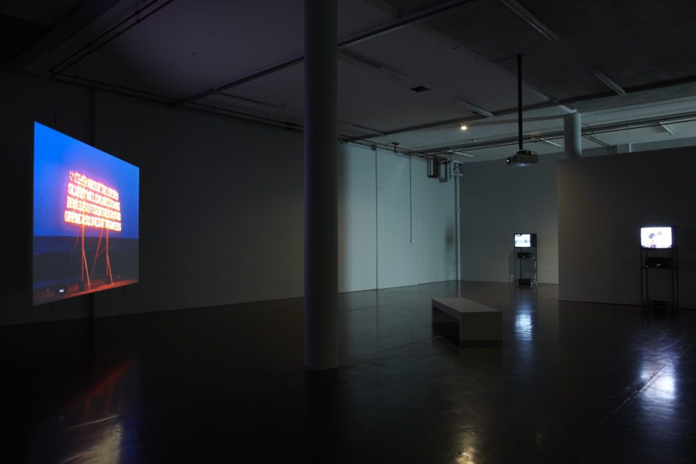 Installation shot: The gallery is dark, three screens show art work.