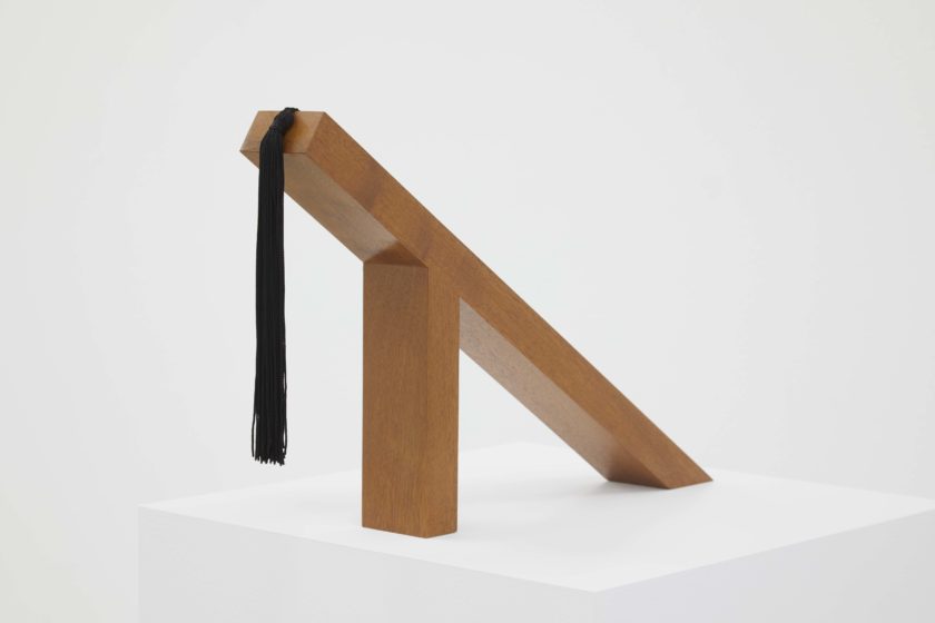 Two pieces of wood have been joined together so one is vertical which supports a piece laying across it diagonally. A black tassle hangs from the top of the diagonal piece.