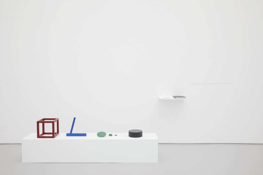 A white plinth on the floor of a gallery has on its surface a red cube outline sculpture, a blue sculpture of a horizontal and angled vertical line, a series of three green sculptures of three different sizes, and a squat black cylindrical sculpture.