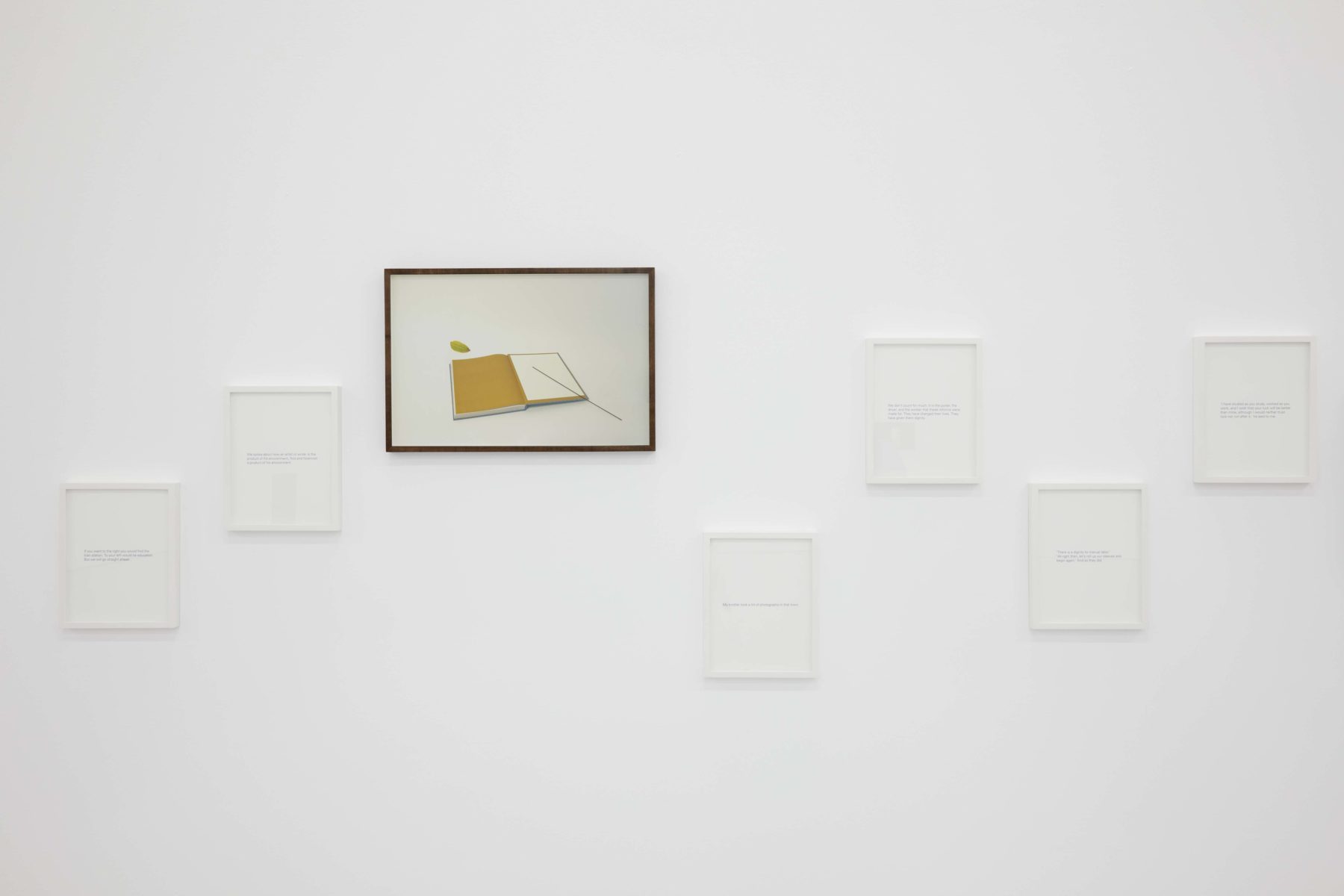 Six white frames hang on the gallery wall. One frame is black and has a photograph of a notebook in it.