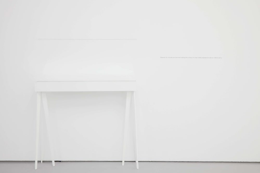 A white desk is placed in front of a white gallery wall so it is only just visible.