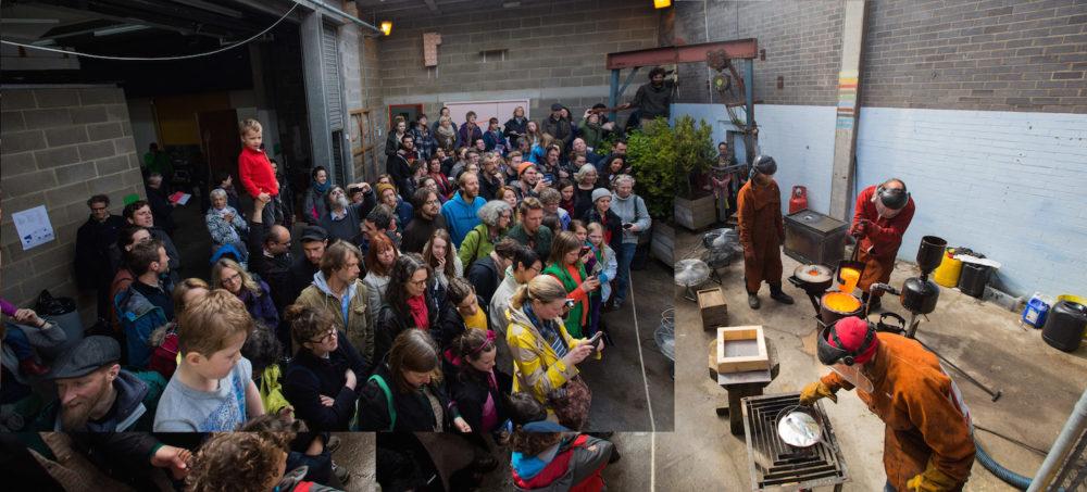 A large group of people are crowded in our sculpture yard watching three people, wearing protective clothing, pour molten metal into a cast.