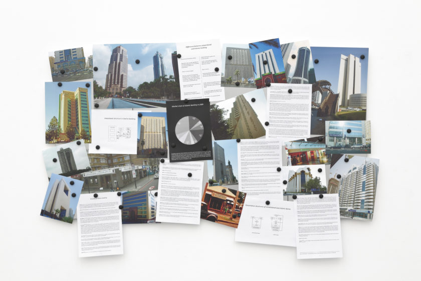 Installation view of Wealth of Nations (2010). Lots of photographs of buildings are pinned to a corkboard, alongside graphs and cards that are heavily printed with text.