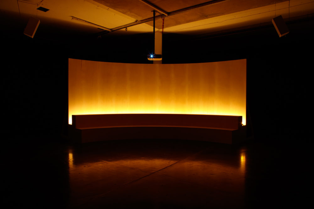 Installation view of Jesse Jones The Struggle Against Ourselves (2012). The seating for the film is a long curved bench and is illuminated in yellow from behind.