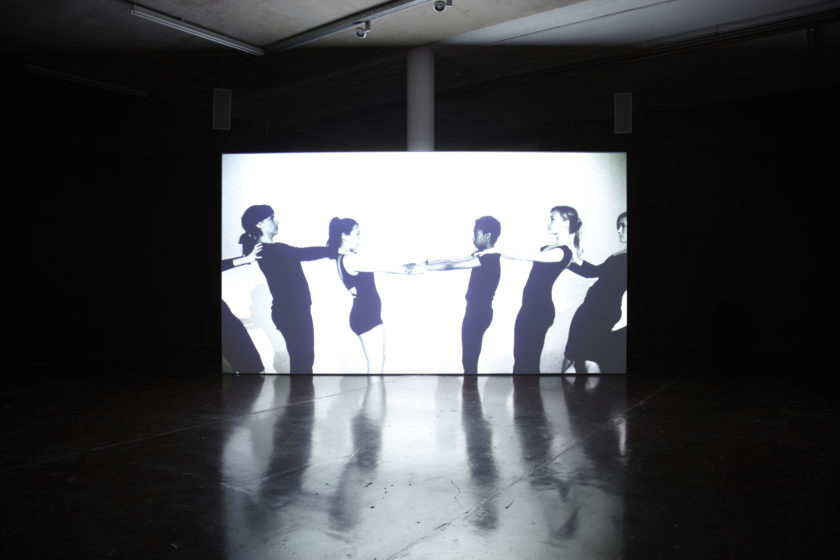 Installation view of Jesse Jones The Struggle Against Ourselves (2012). A projection of five people with their hands on one anothers' shoulders plays in a dimly lit gallery.