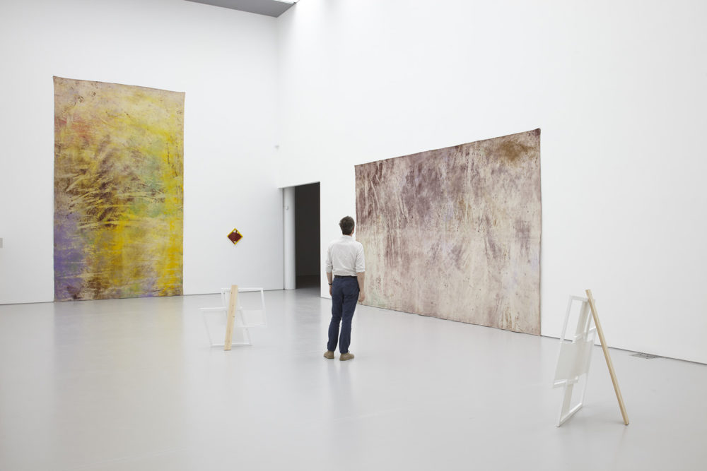 Installation view of Jessica Warboys, Ab Ovo (2013). A visitor studies large scale pastel textiles hung on the wall. The gallery is light and painted white.