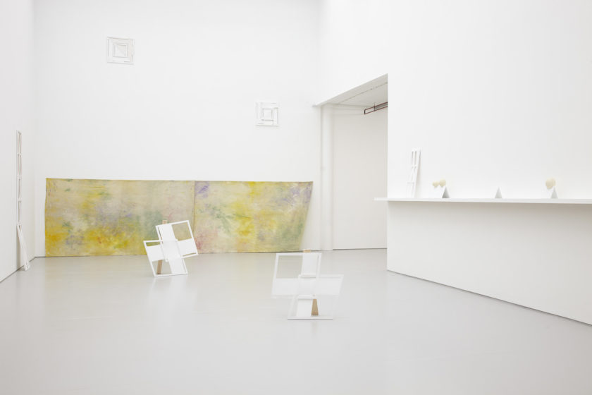 Installation view of Jessica Warboys, Ab Ovo (2013). The gallery is light and painted white. Multi-coloured pastel sheets hang on the faraway wall. White structures sit on the floor in the foreground.