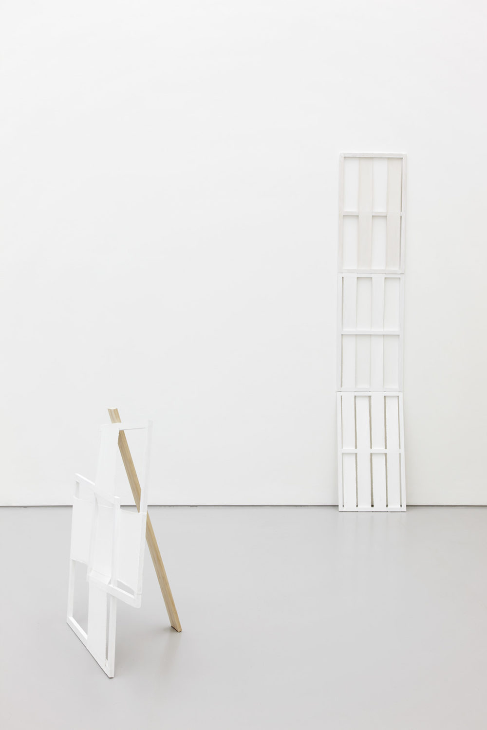 Installation view of Jessica Warboys, Ab Ovo (2013). The gallery is light and painted white. One long white geometric structure leans against a wall, another sits in the foreground leaning against a plank of wood - together they make an 'A frame'.