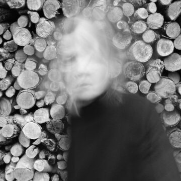 Black and White photograph of artist Kamina Walton in motion in front of wall of logs.