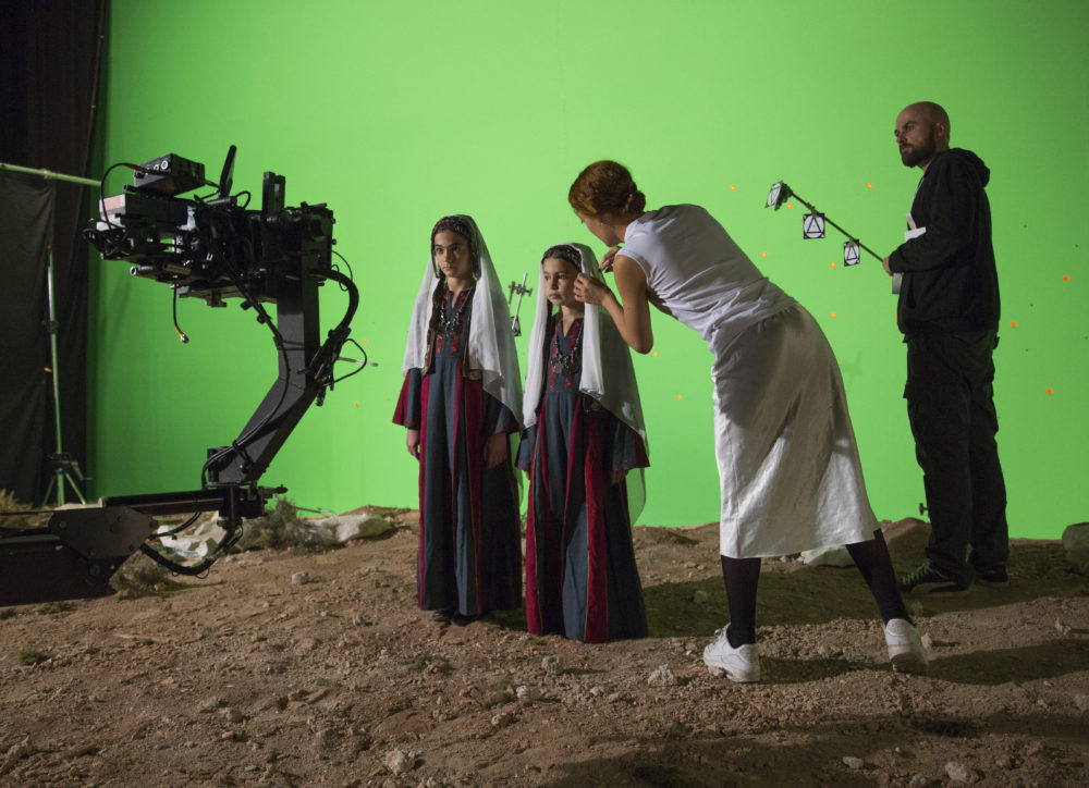 Production shot: Two young people are stood on sand in front of a green screen, they are wearing white headscarfs whilst the artist tends to their clothes. A member of the crew stands behind.