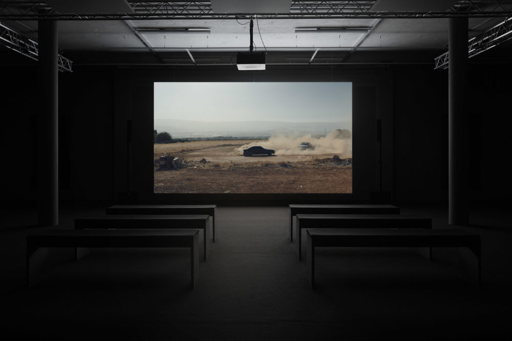 Installation view: A screen shows a car surrounded by dust it has made. In front of the screen are six benches.