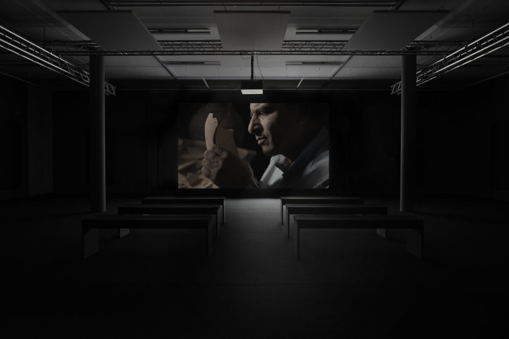 Installation view: A screen shows a man sticking broken pottery back together.