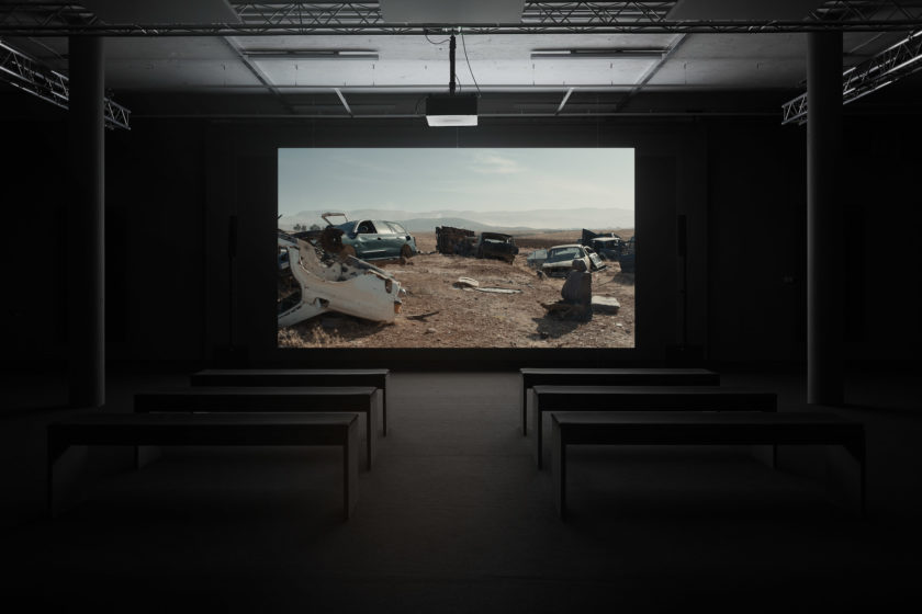 Installation view: A screen shows cars in a junkyard in a sandy environment.