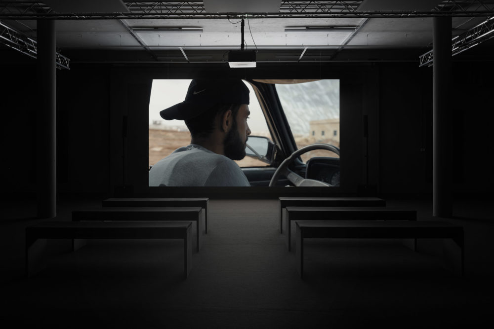 Installation view: A screen shows a man wearing a backwards cap driving a car.