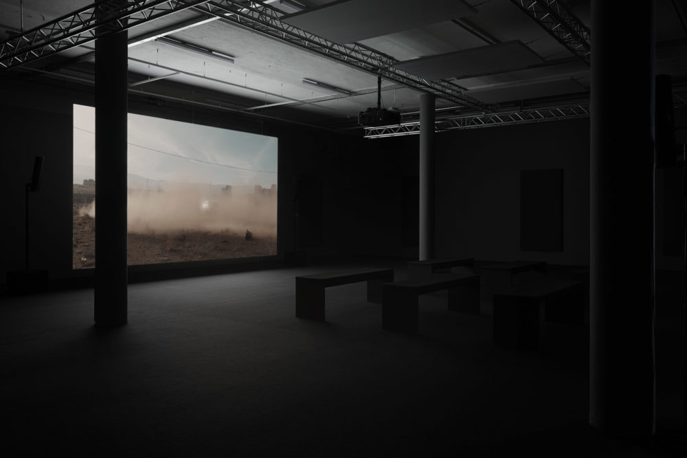 Installation view: A screen shows a car surrounded by dust it has made. In front of the screen are six benches.