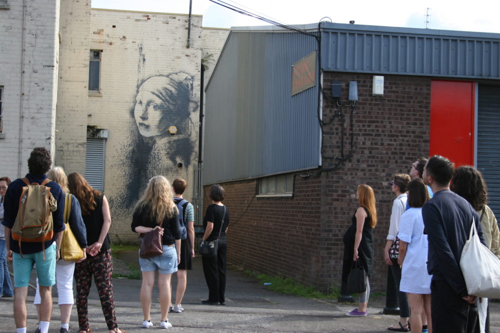A group of people stand in a car park. A famous mural by Banksy, The Girl With the Pierced Eardrum, is on the wall behind them.