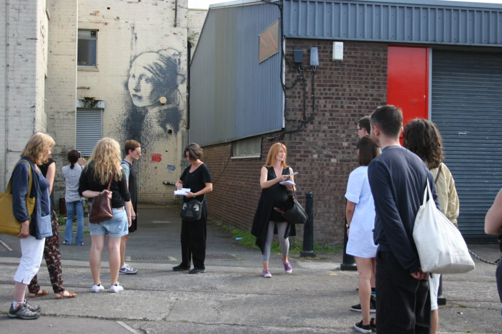 A group of people stand in a car park. A famous mural by Banksy, The Girl With the Pierced Eardrum, is on the wall behind them.
