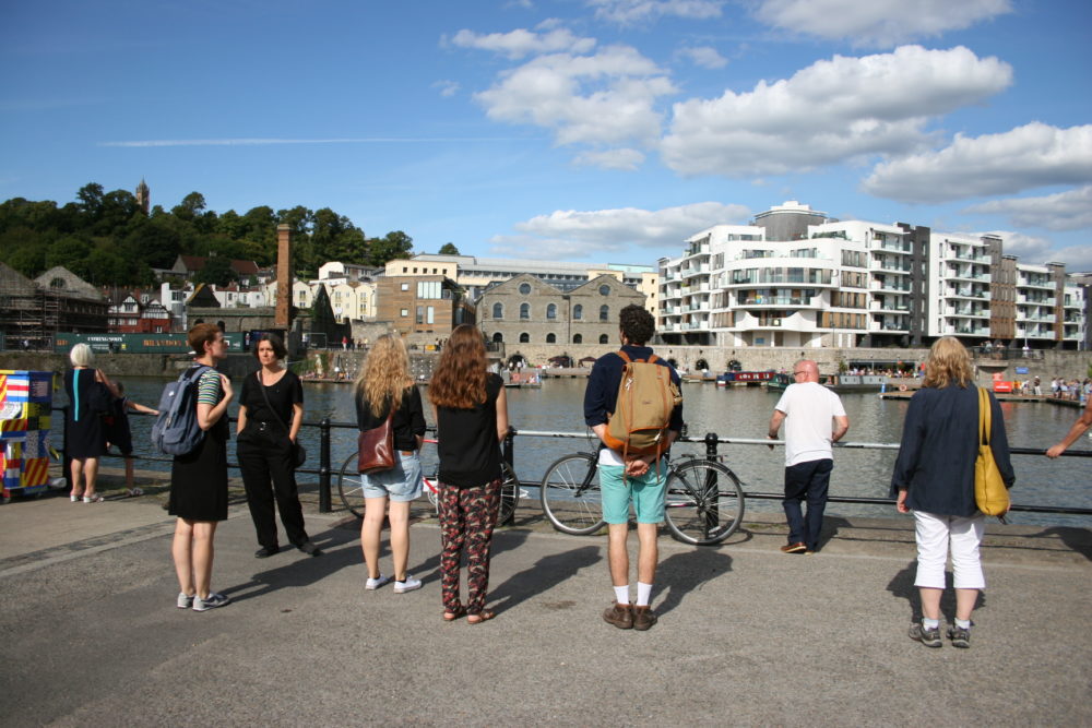 A group of people look out at Bristol Harbour on a sunny day.