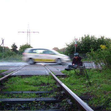 A photograph of a man using recording equipment next to a level crossing. A car crosses the level crossing in a blur.