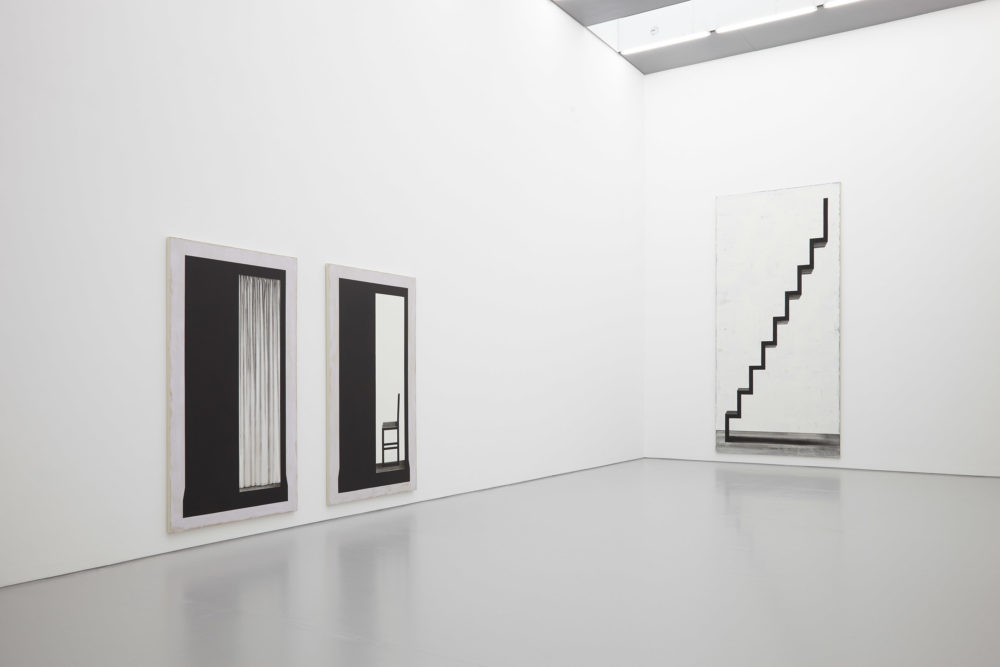 Installation shot: Three large black and white paintings hang in the gallery. Two are of a photo booth, and the other is of a staircase.