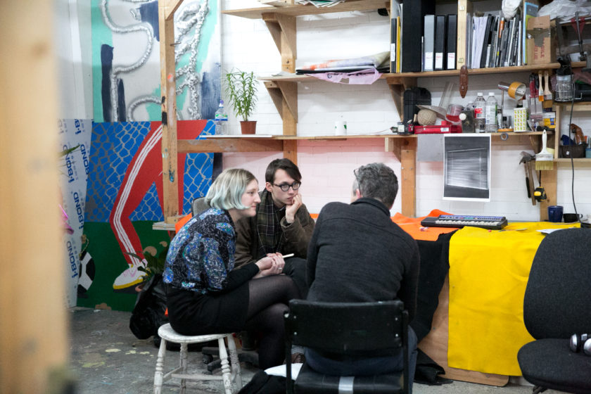 Three people sit together having a meeting in an artists' studio.