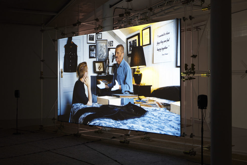 The screen shows a scene of two men sat in deep discussion in a bedroom. A pair of leather trousers are laid on the bed, as are lots of boxes.