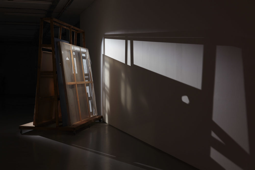 A large wooden structure with building materials stacked on it casts a dramatic shadow in the gallery.