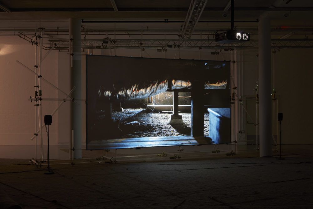 The scaffolding supported screen shows a scene of a basement beneath a working building - a thick layer of dust lies on the floor and a large ceiling pipe is covered in foil.