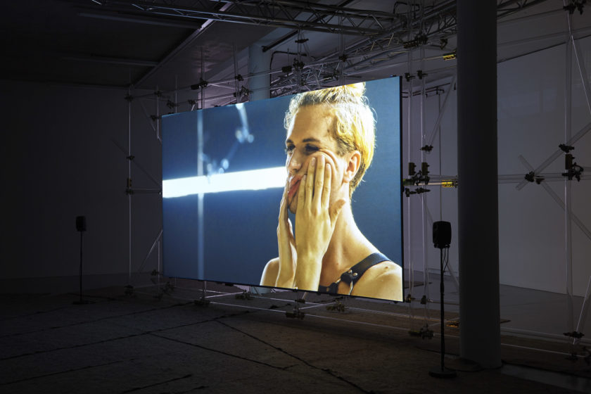 A screen has been erected at Spike Island gallery using a lot of scaffolding. On the screen, a blonde person is wearing leather bondage gear and kneading their face.