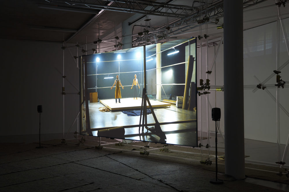 The scaffolding supported screen shows a platform with two men stood on it with their arms outstretched.