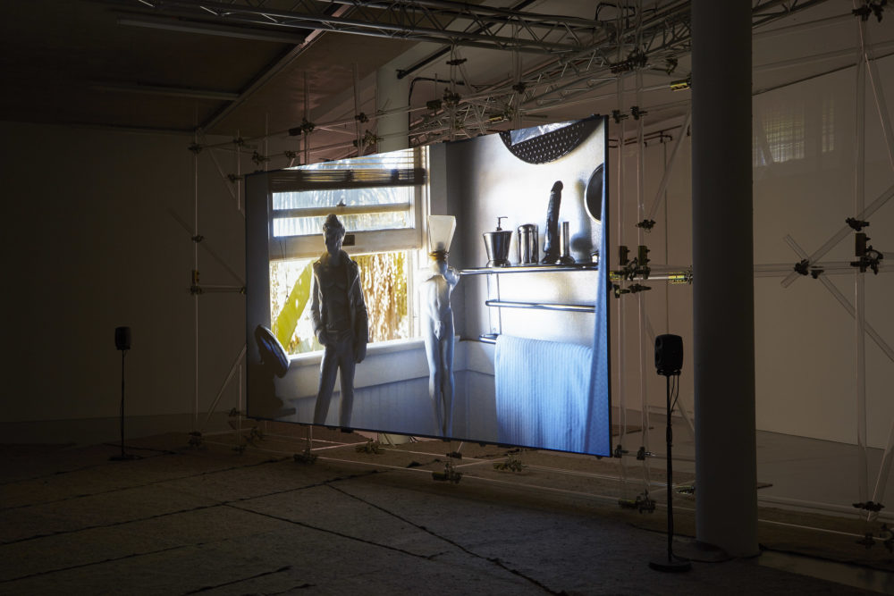 The scaffolding supported screen shows a bathroom window. In the bathroom you can see two classic greek sculptures, a towel rack and a large dildo on a shelf.